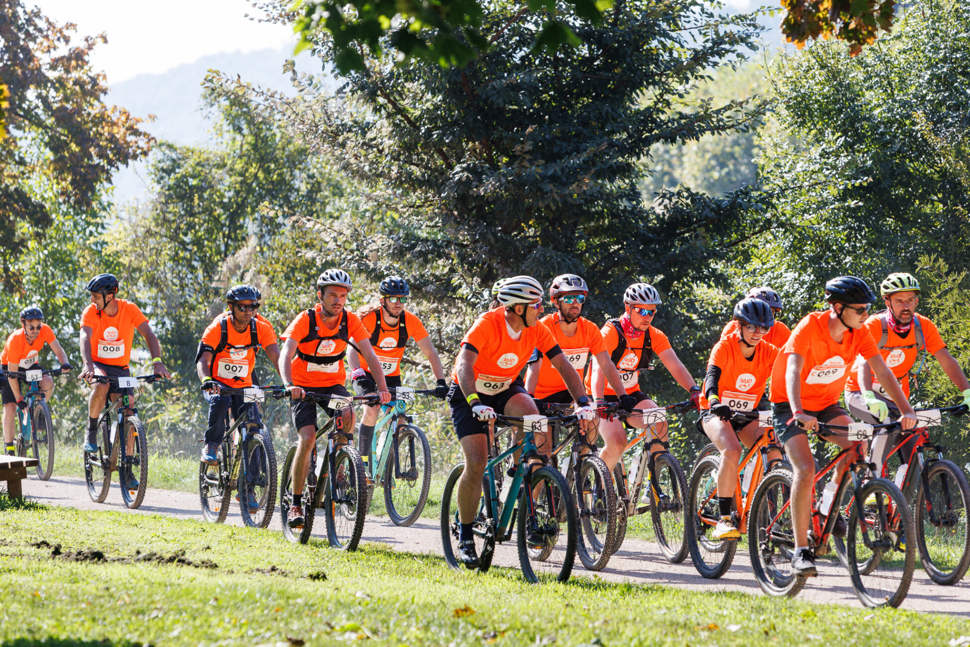 Group of cyclists wearing matching orange shirts and helmets riding mountain bikes on a sunny trail