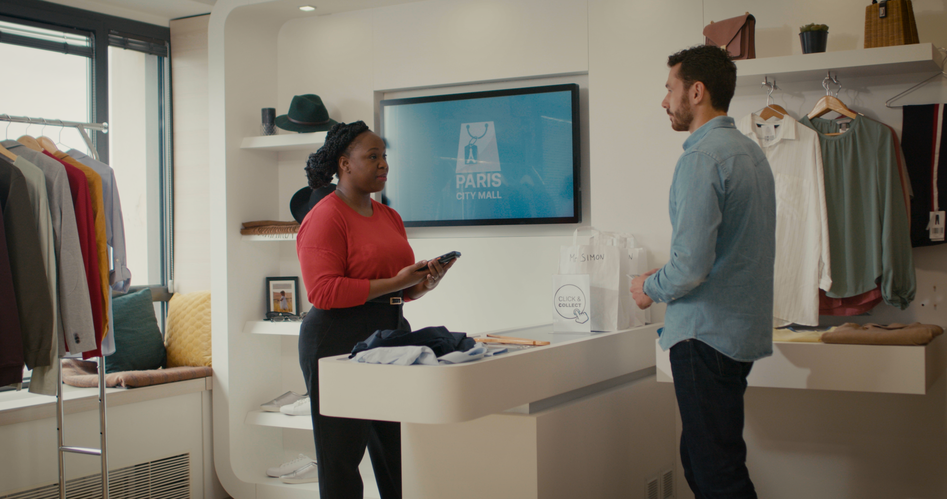 Boutique staff assisting customer at checkout counter with Click & Collect bags, Paris City Mall screen in background