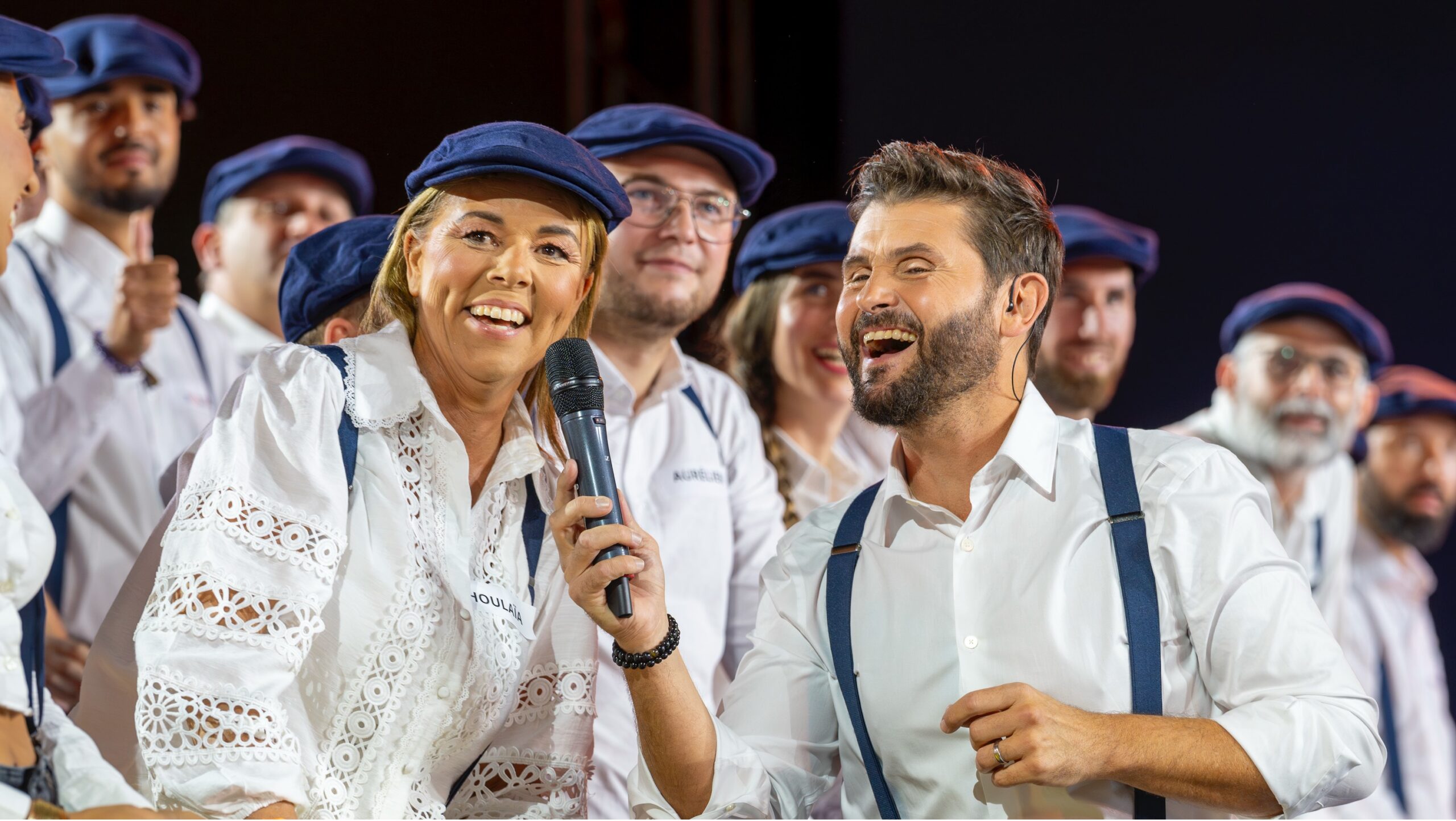 Group of people dressed in white shirts and blue caps, man holding microphone laughing next to smiling woman