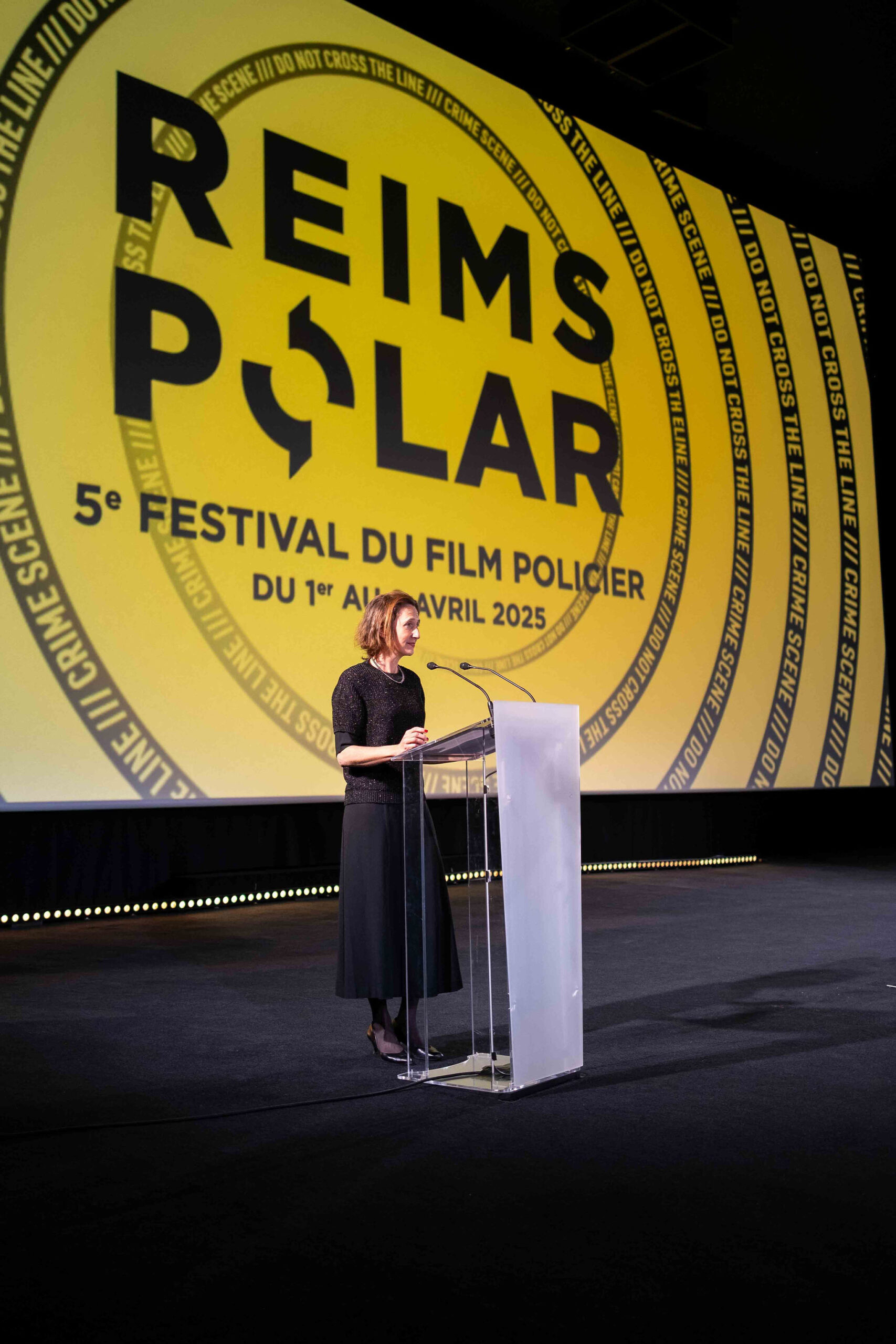 Woman speaking at podium during Reims Polar Crime Film Festival with large yellow event backdrop