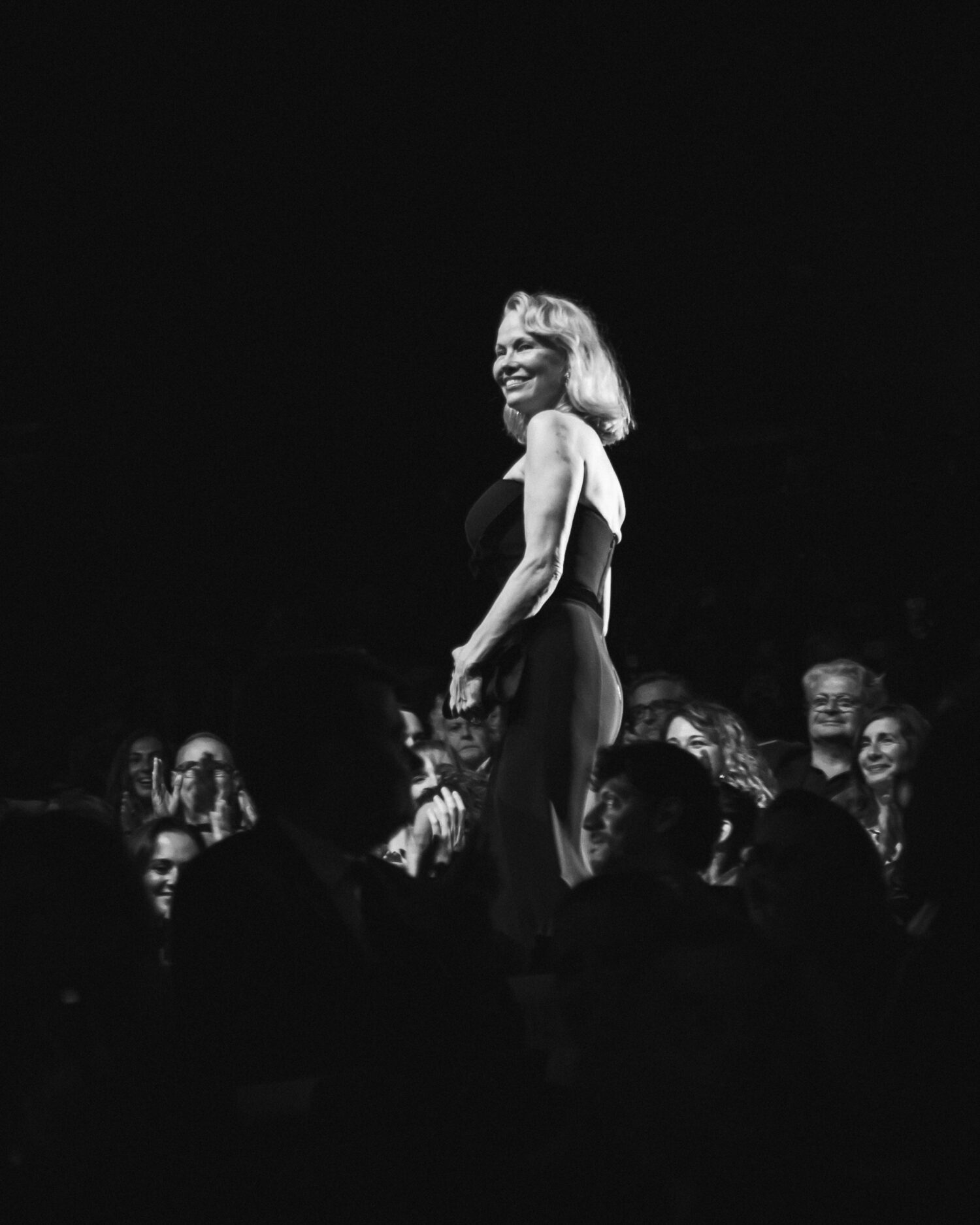 Smiling woman in elegant dress standing and applauded by audience in dimly lit theater
