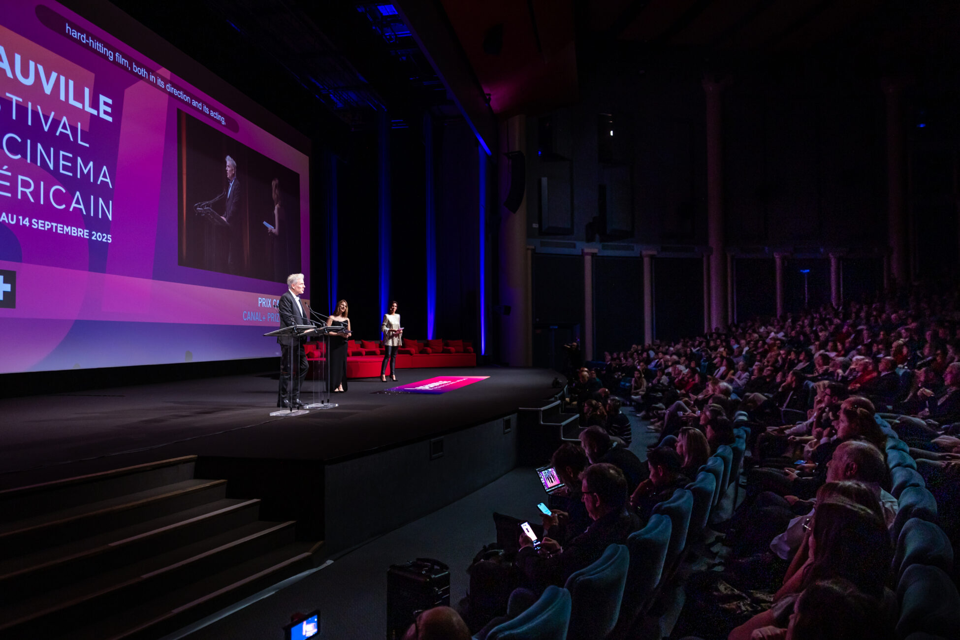 Speaker at podium addressing large audience during Deauville American Film Festival event in theater