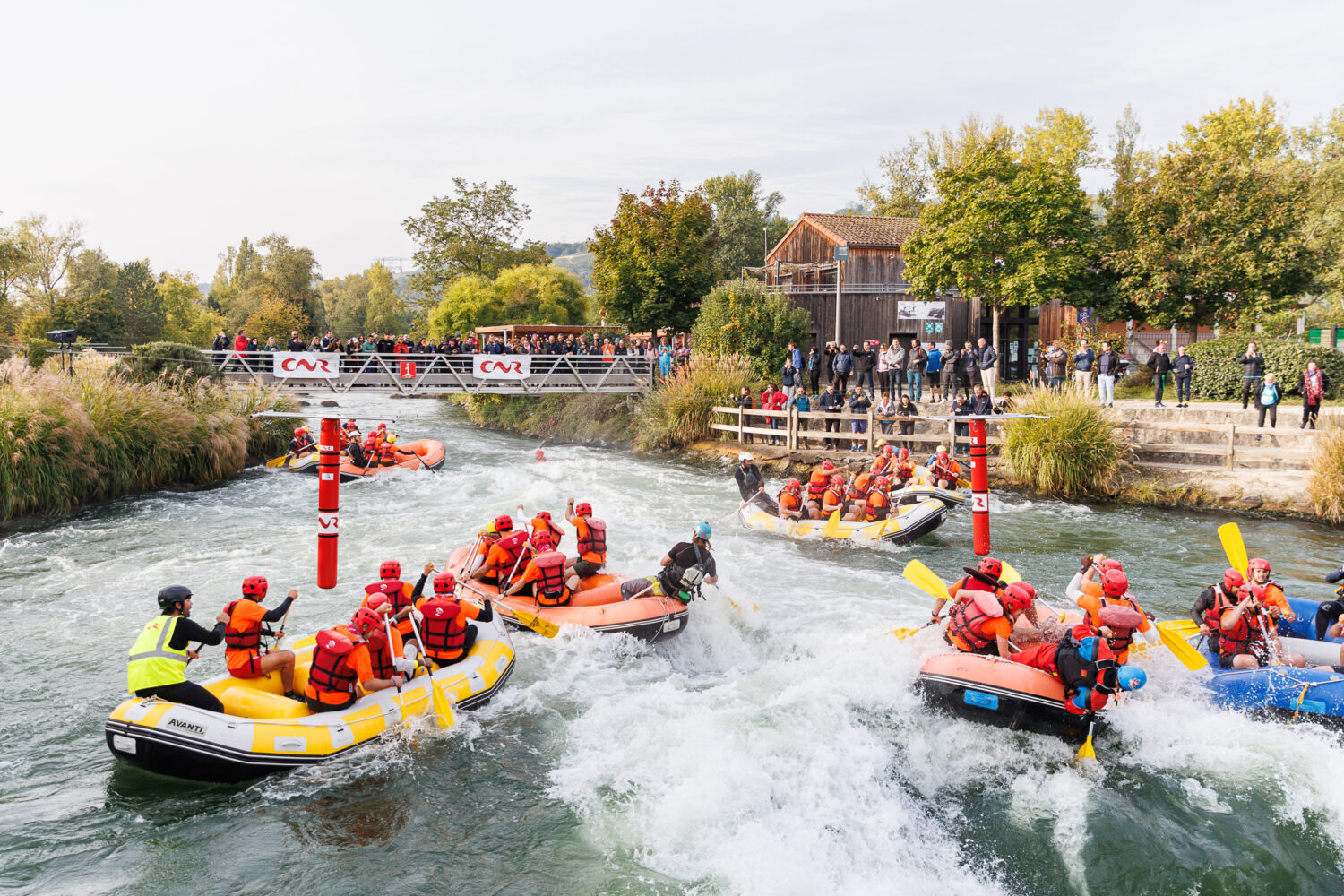 Multiple rafts with people in orange life jackets navigating fast-moving river with spectators on banks and bridge