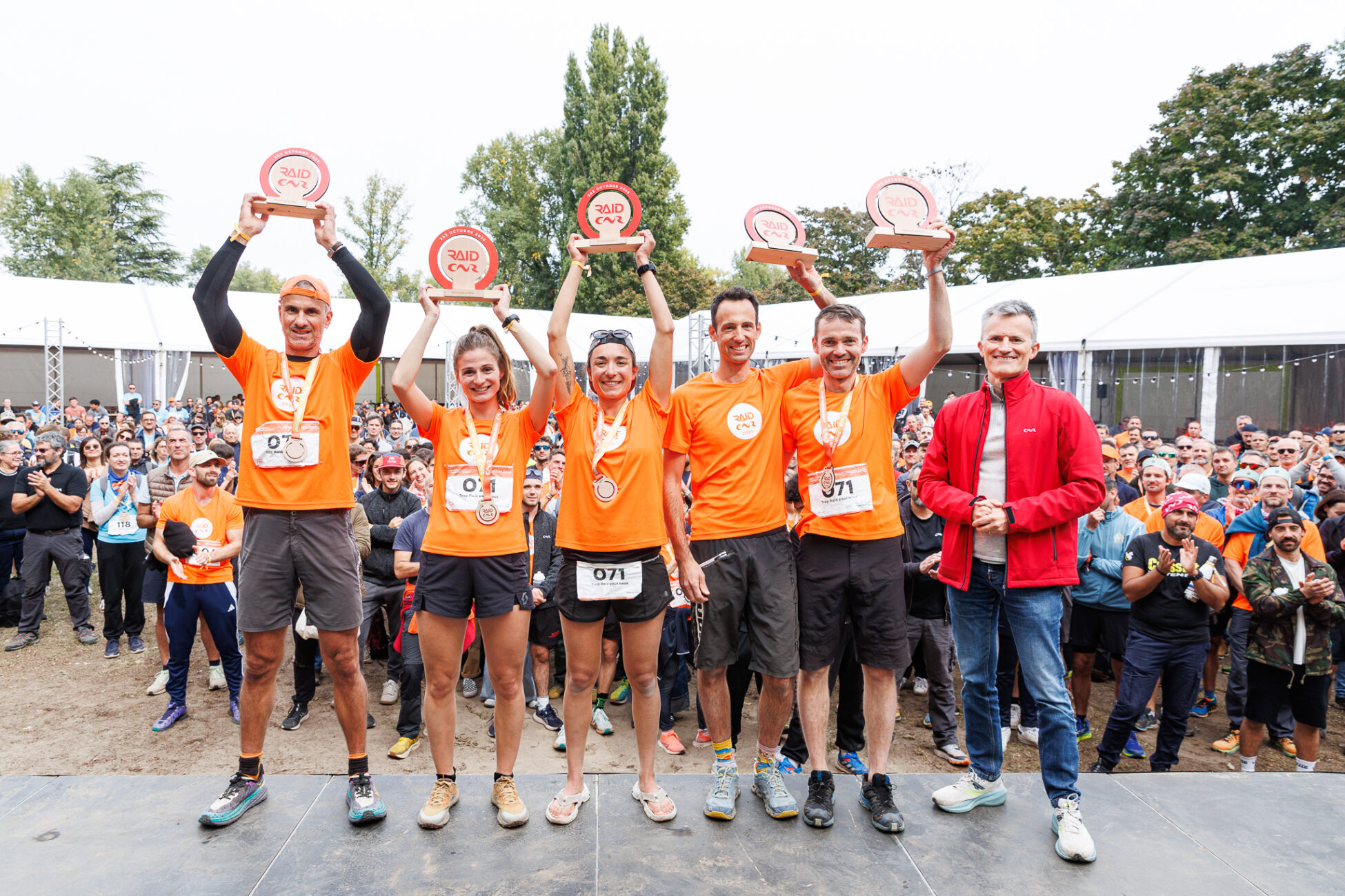 Group of athletes in orange shirts holding up trophies on stage at outdoor sports event with crowd cheering