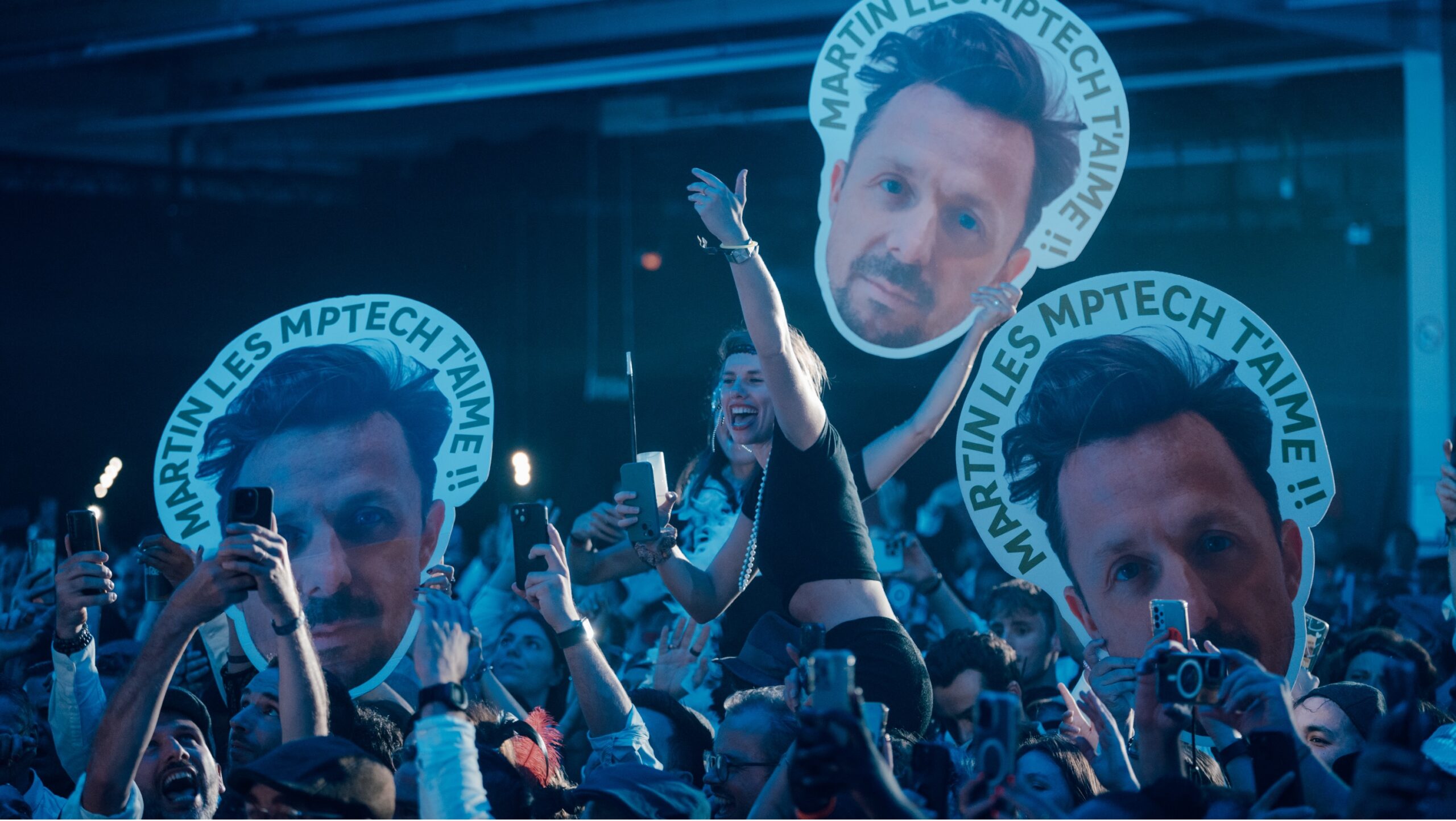 Excited crowd holding large cutouts of a man's face, some taking photos with smartphones at a lively event