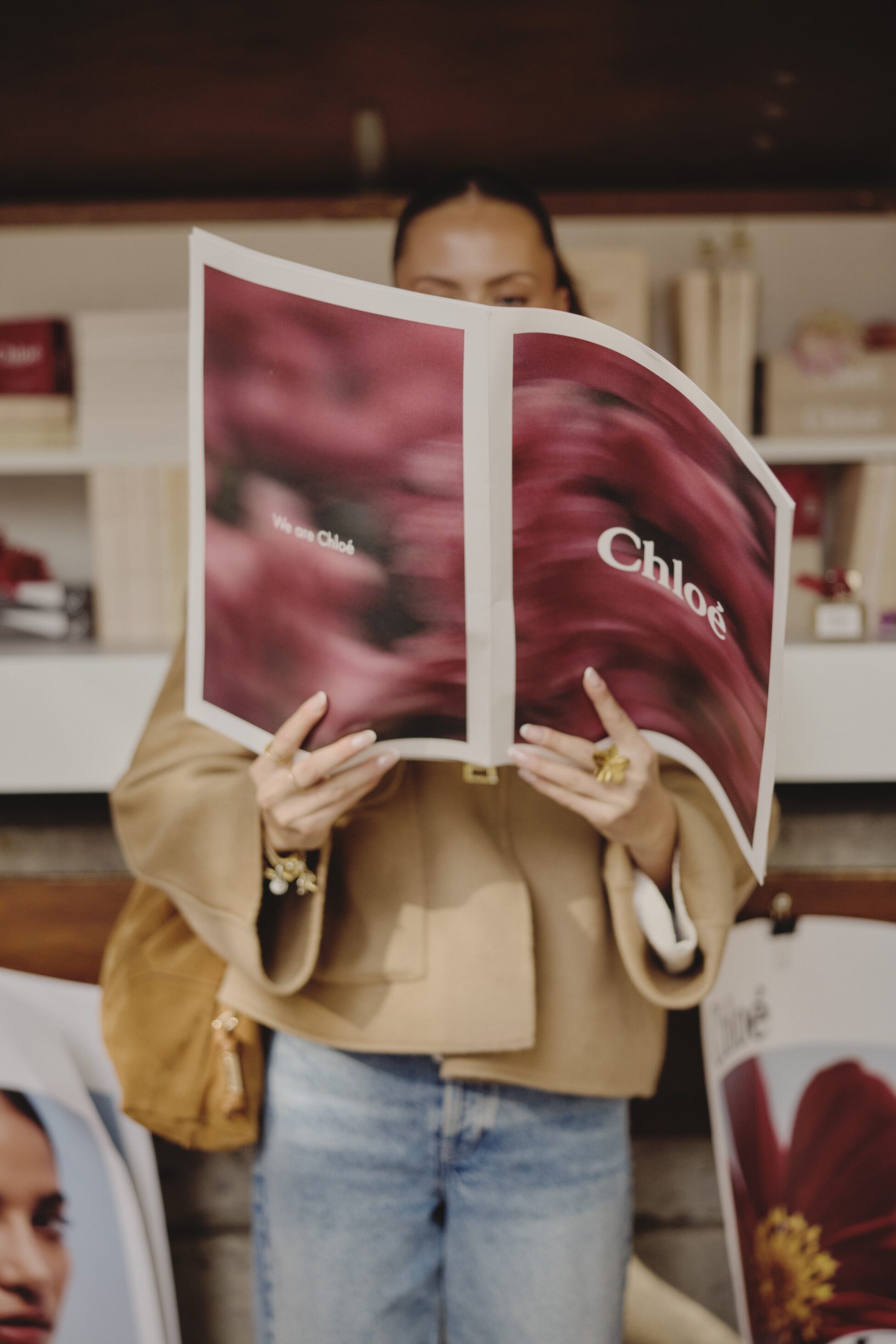 Woman holding a large Chloé branded magazine with blurred floral cover wearing beige jacket and blue jeans