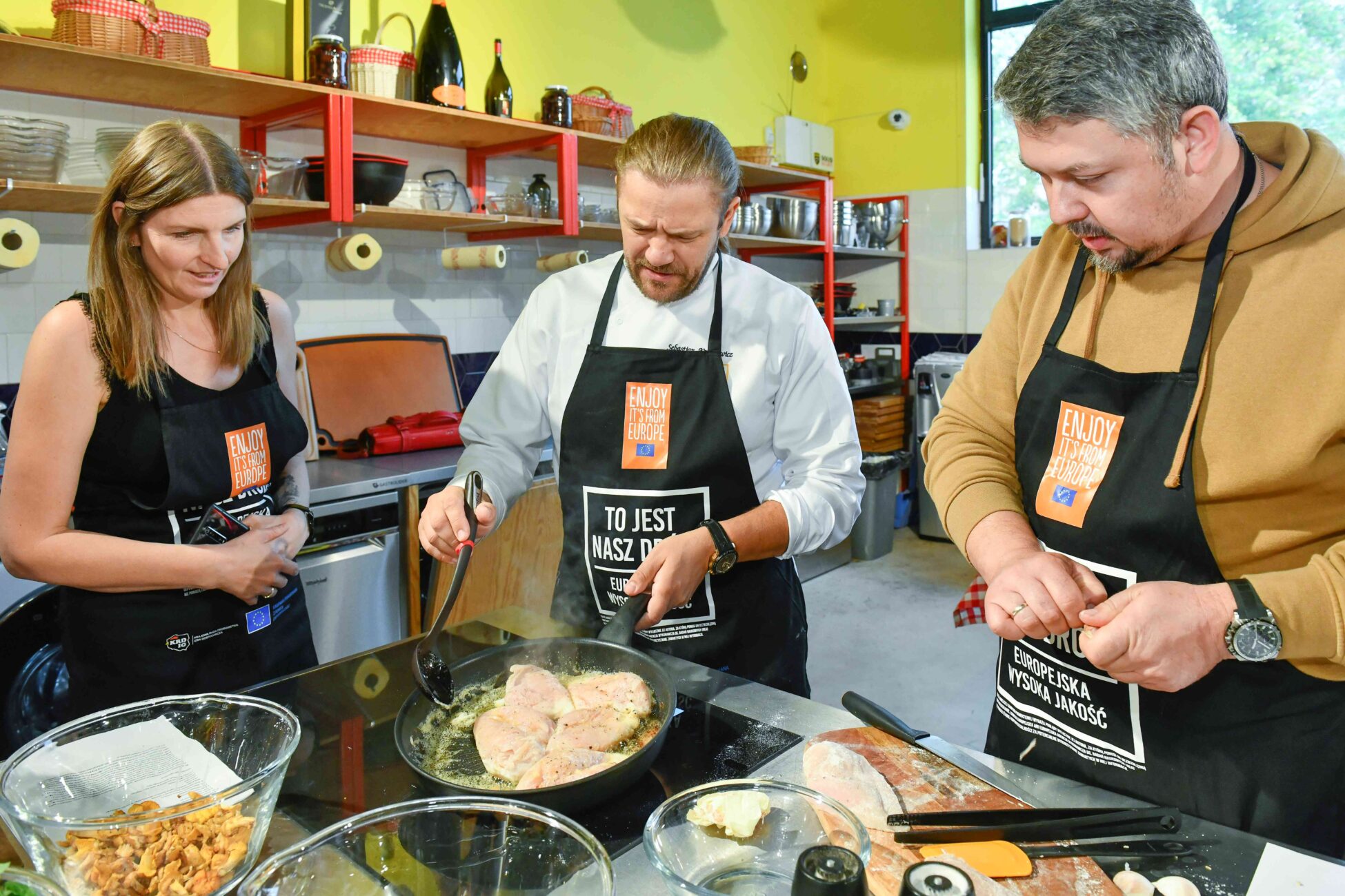 Chef leading a cooking class searing chicken breasts in a skillet with two participants, all wearing “Enjoy It’s From Europe” aprons in a professional kitchen